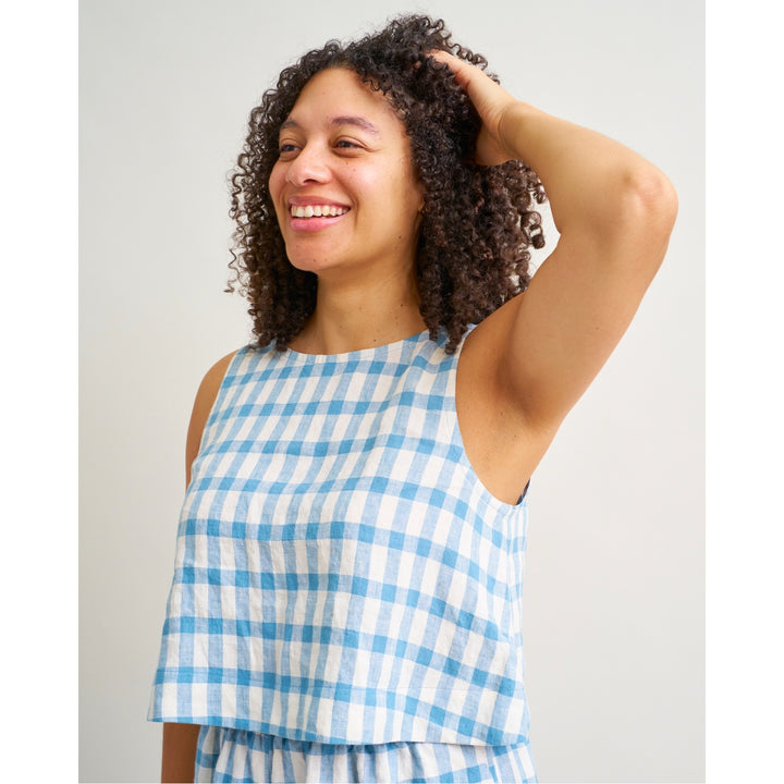 A woman with curly brown hair smiling and looking to the side, wearing a sleeveless blue and white gingham check top against a plain light gray background.