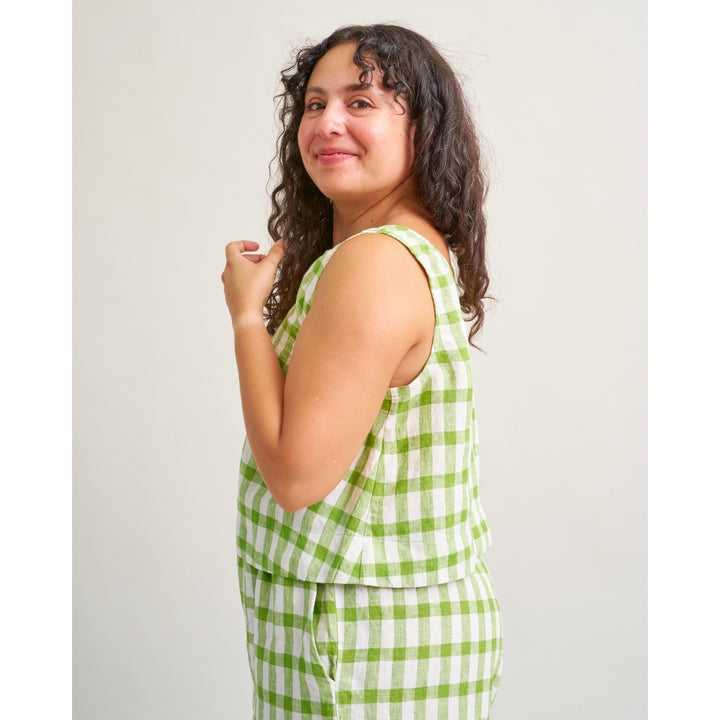 A woman with curly dark hair is shown from the side wearing a sleeveless green and white gingham check top and matching bottoms against a plain light gray studio background.