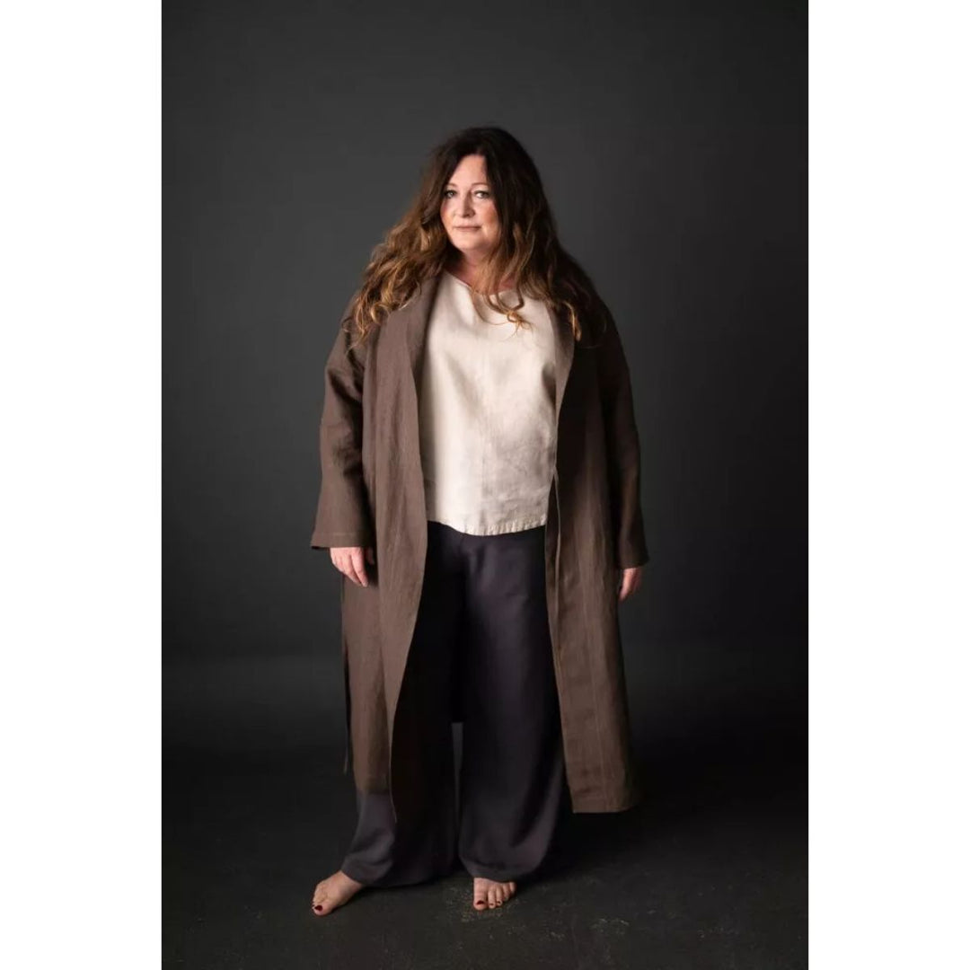 Woman wearing a long brown coat over a white sweater and dark pants on a black background