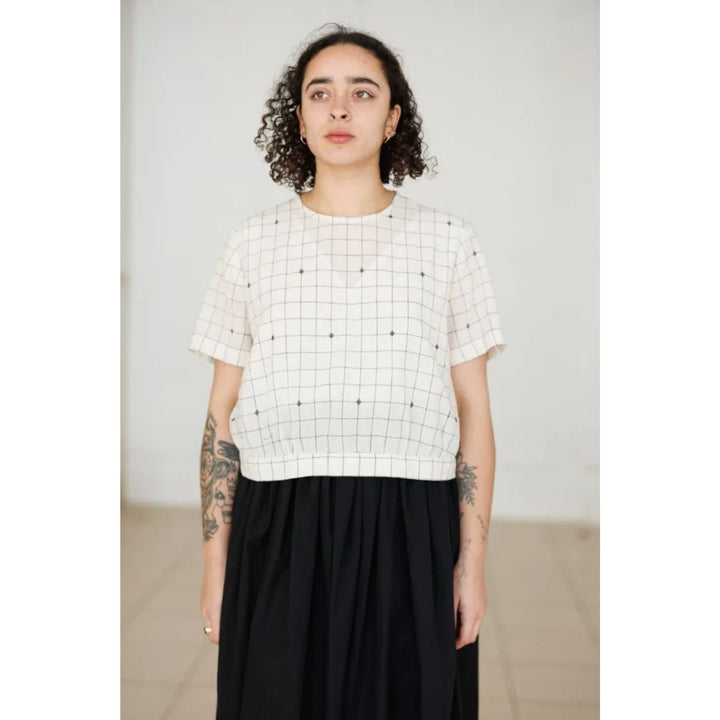 Person wearing a white grid-patterned top and black skirt against a plain background