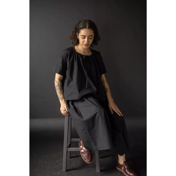 Woman sitting on a stool wearing a black outfit against a dark background