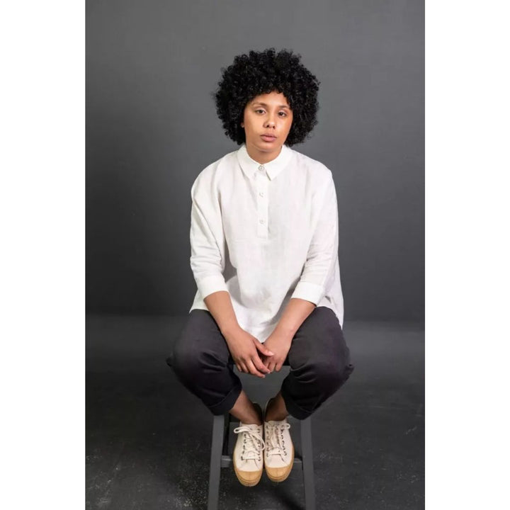 Person wearing a white shirt and dark pants sitting on a stool against a gray background