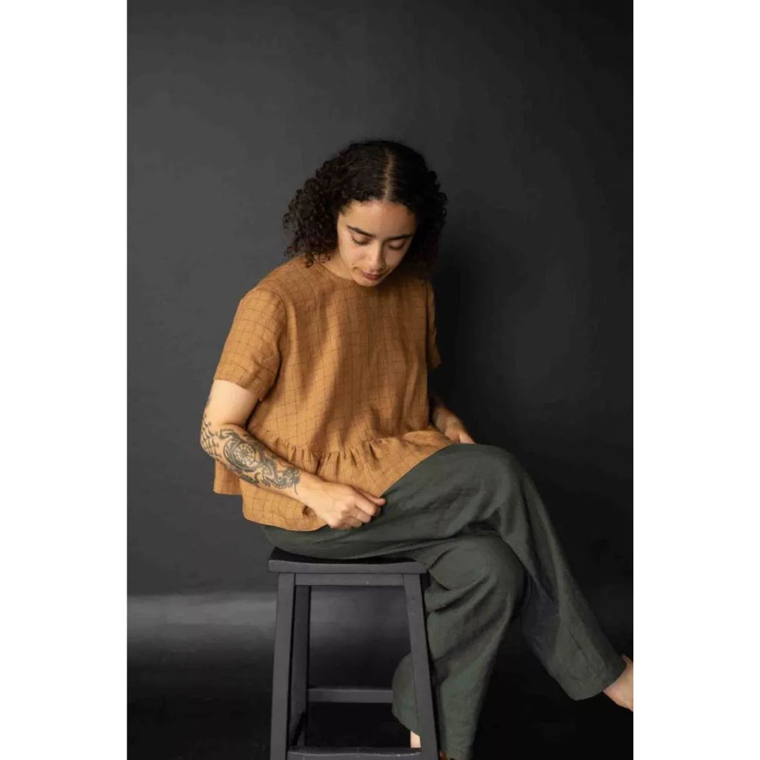 Person wearing a brown top and green pants sitting on a black stool against a dark background