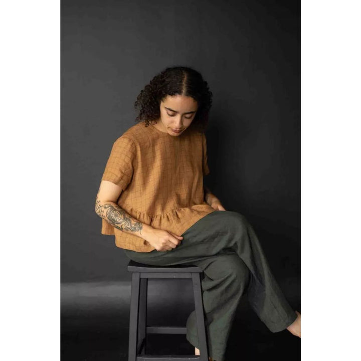 Person wearing a brown top and green pants sitting on a black stool against a dark background