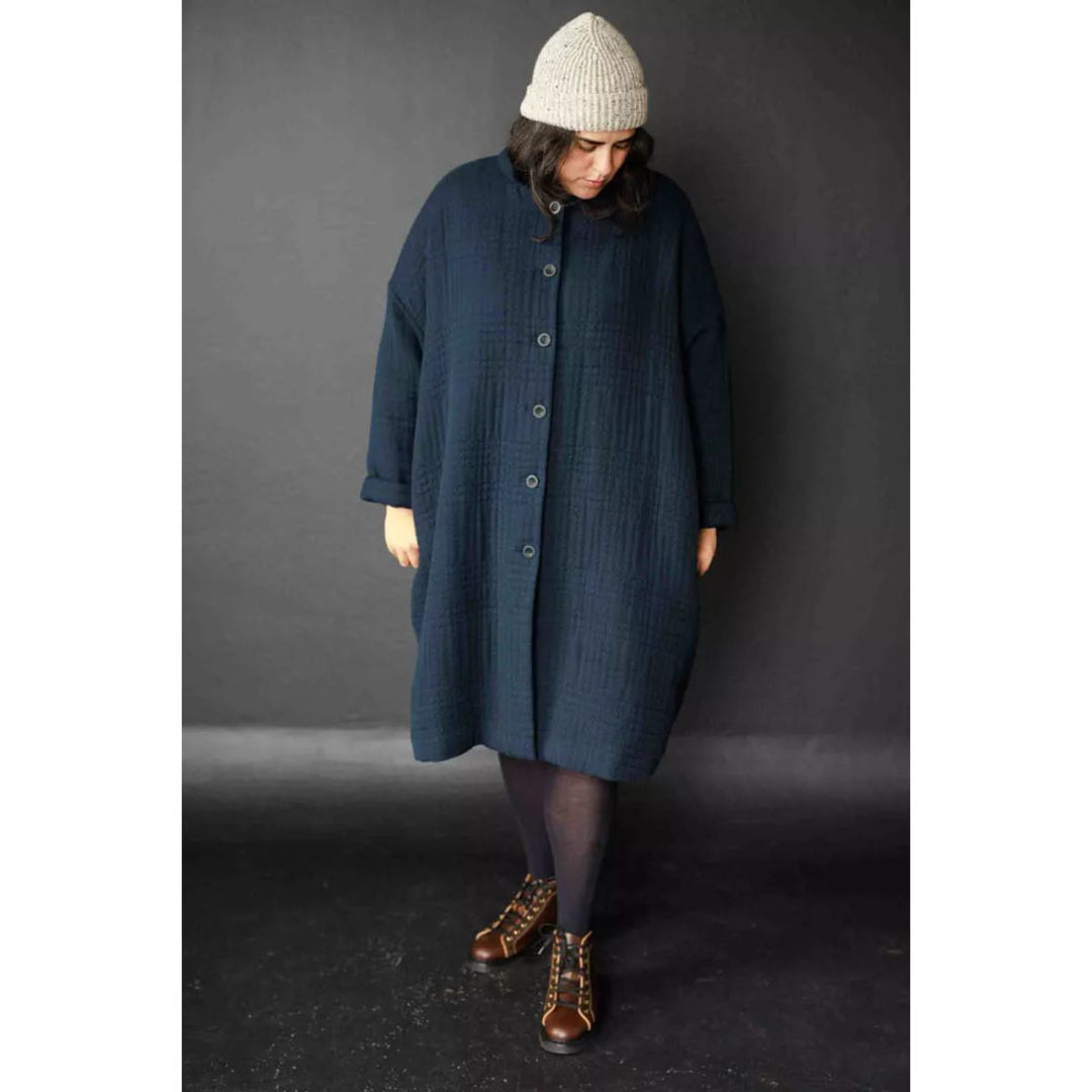 Person wearing a long dark blue coat and a white knit hat against a gray background