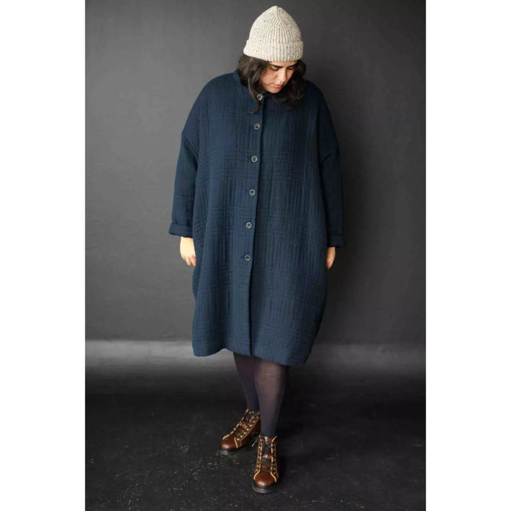 Person wearing a long dark blue coat and a white knit hat against a gray background