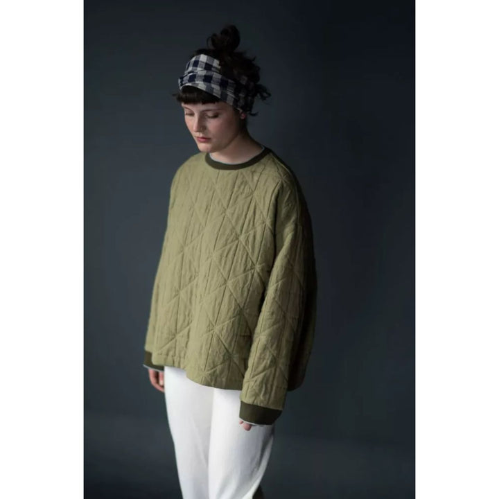 Person wearing a quilted green sweater and white pants against a dark background