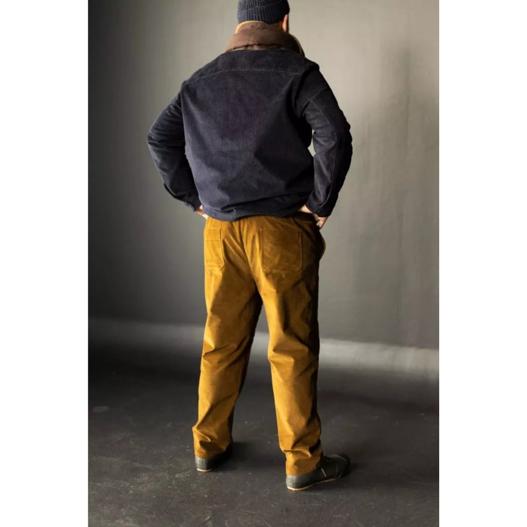 Person wearing a dark jacket and mustard pants standing against a plain wall.