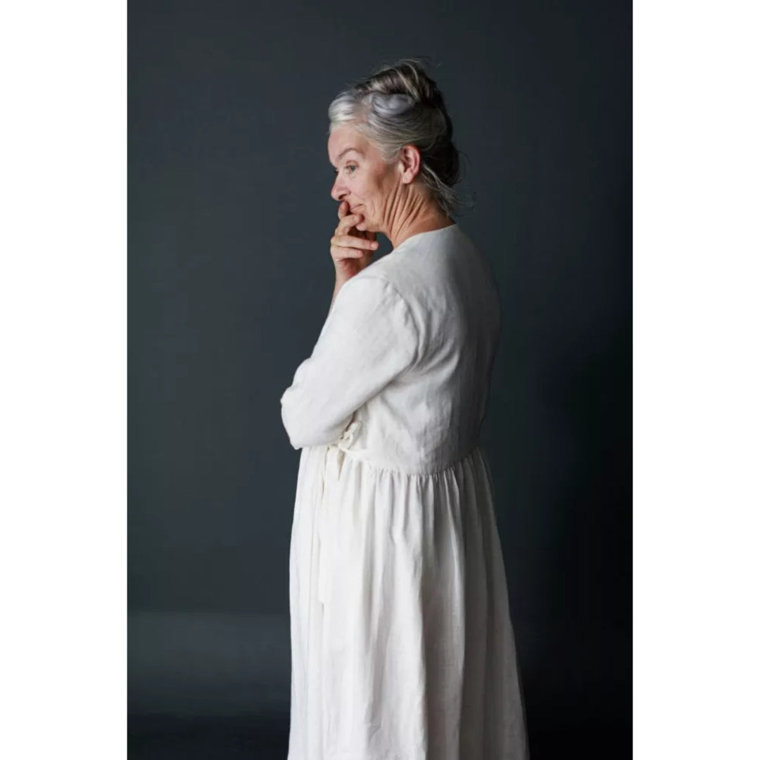 A woman with gray hair in a bun wearing a long-sleeved white linen dress, shown from a side-back profile against a dark gray studio background.