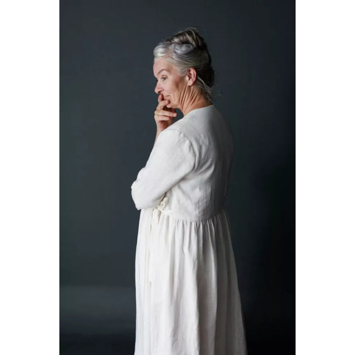 A woman with gray hair in a bun wearing a long-sleeved white linen dress, shown from a side-back profile against a dark gray studio background.