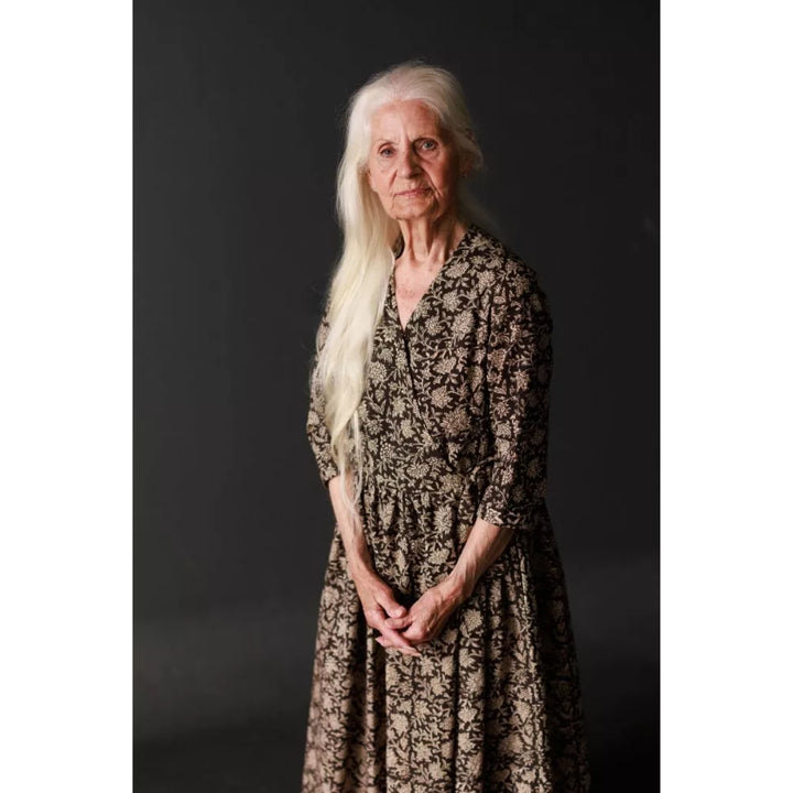 An elderly woman with long white hair stands against a dark studio background wearing a long-sleeved, knee-length dress with a dark brown floral pattern on a beige base.