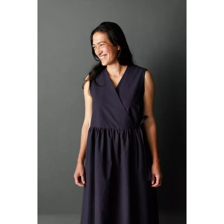 A woman wearing a sleeveless wrap-style midi dress in a deep navy blue, standing against a dark gray studio background.