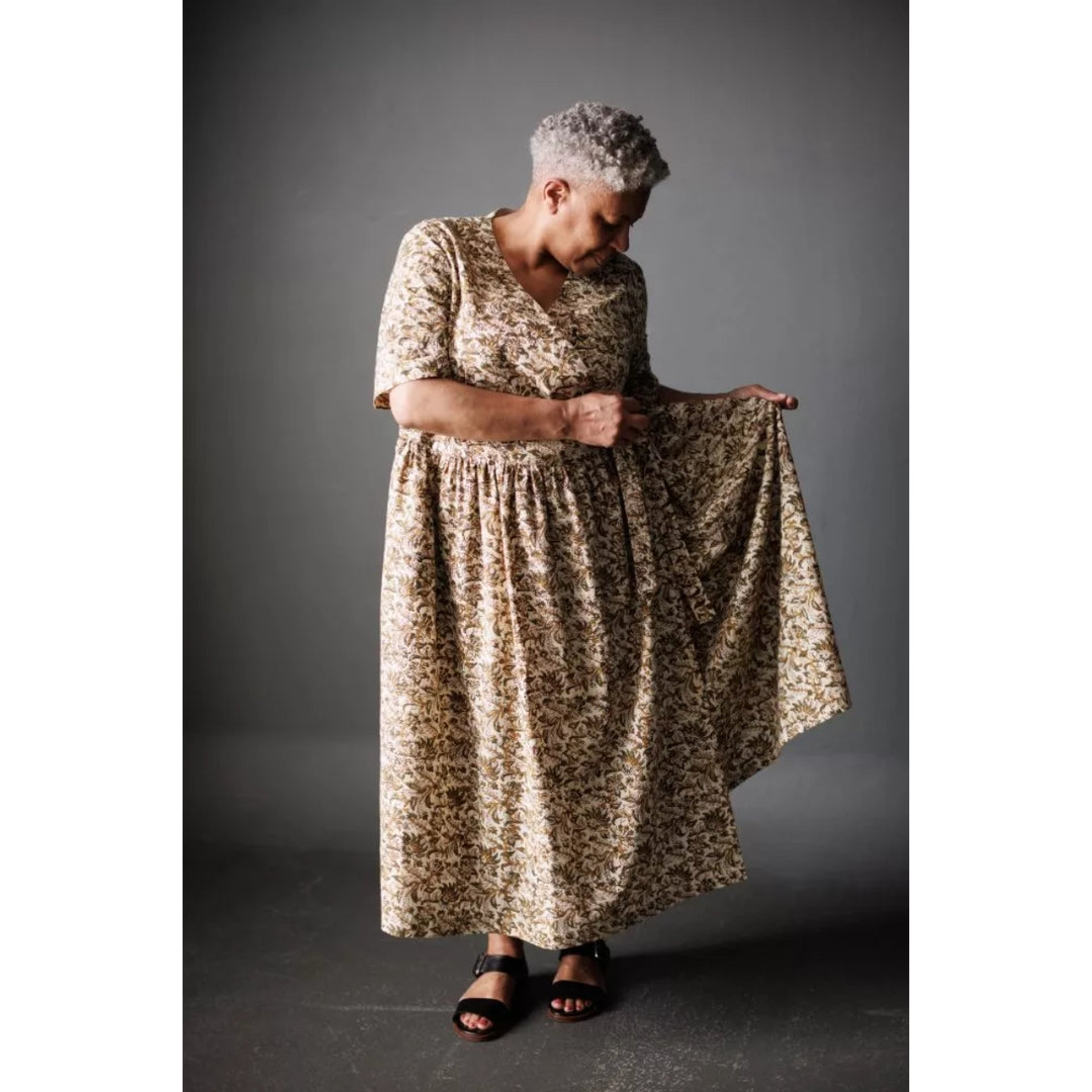 A woman with short gray hair models a knee-length dress with a beige and brown floral pattern, holding out the skirt to show its flow and drape against a dark gray studio background.