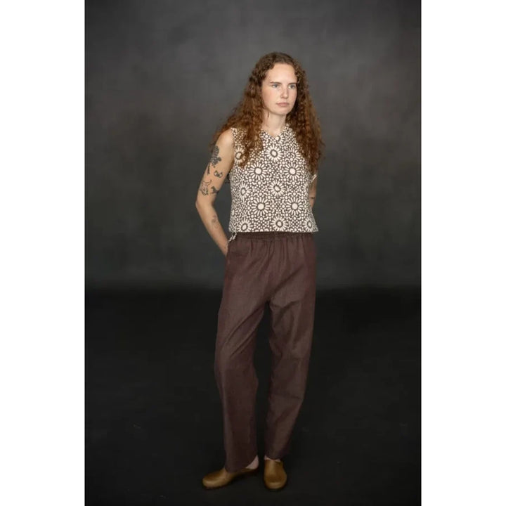 A model with long curly brown hair stands against a dark gray studio backdrop, wearing a sleeveless cream top with a brown geometric pattern and loose brown pants.