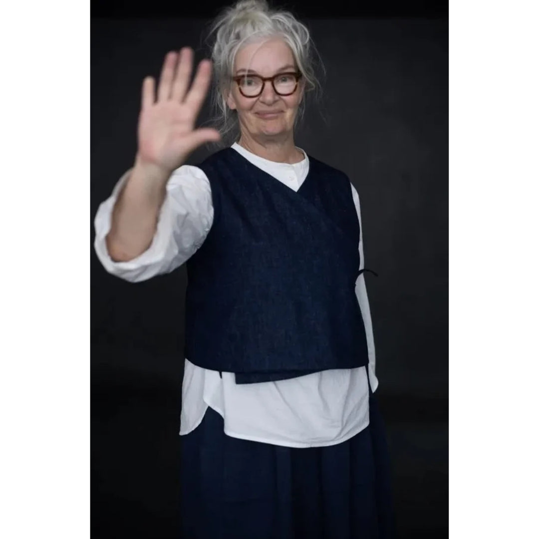 A person with gray hair and glasses wearing a dark navy vest over a white shirt, posing with one hand raised against a dark studio background.