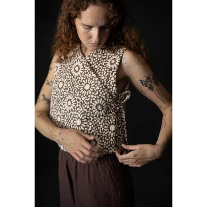 A woman with curly brown hair wearing a sleeveless wrap-style top with a brown and beige floral pattern, paired with dark brown pants, posed against a solid black background.
