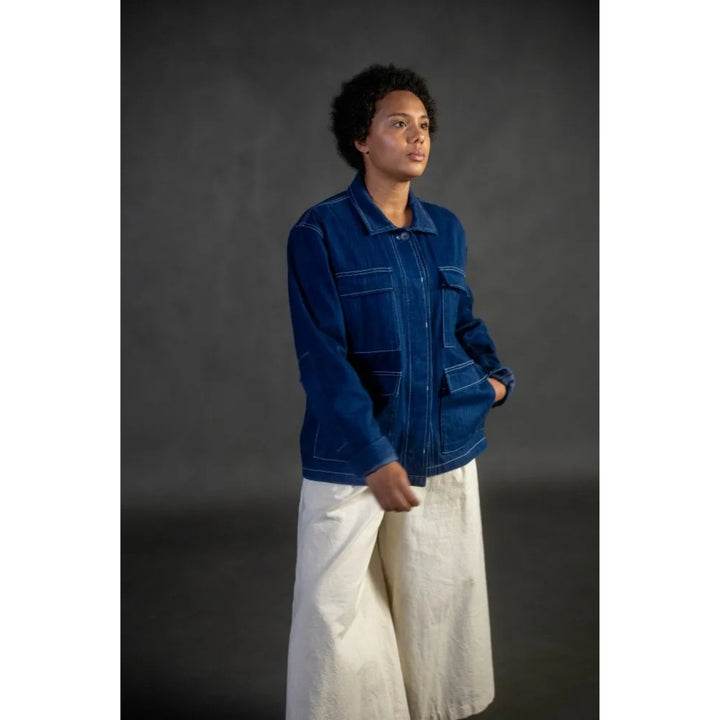 A model wearing a dark blue denim jacket and off-white wide-leg trousers, standing against a dark gray studio background.