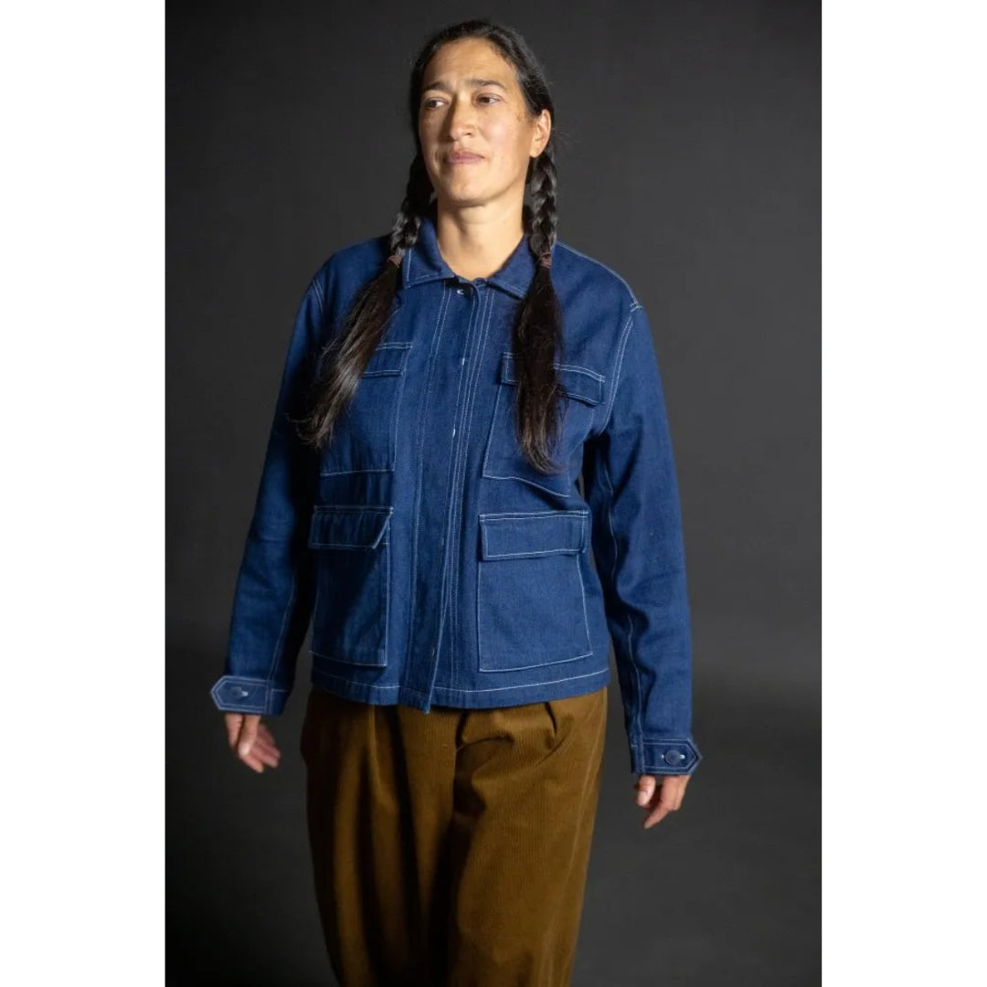 A model with long dark braids wearing a blue denim jacket with contrast white stitching and brown pants, standing against a dark gray studio background.