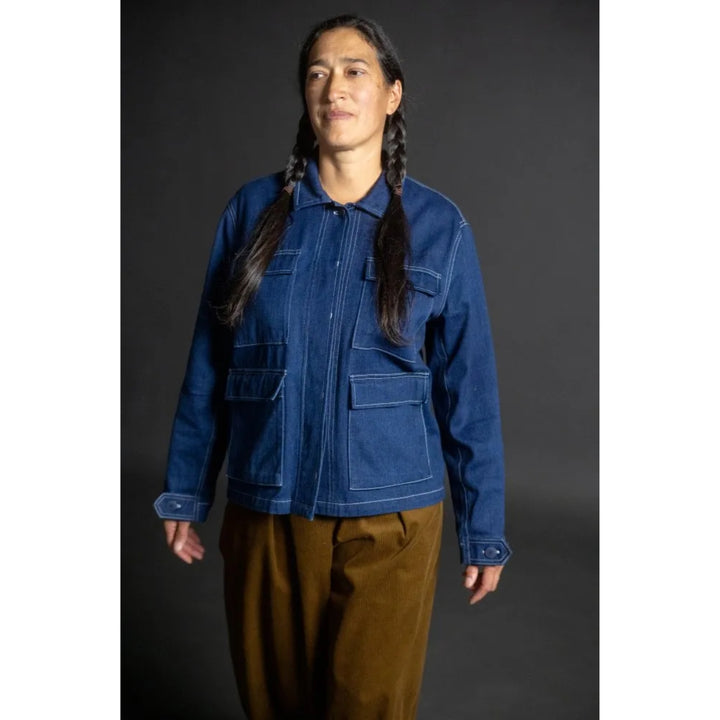 A model with long dark braids wearing a blue denim jacket with contrast white stitching and brown pants, standing against a dark gray studio background.