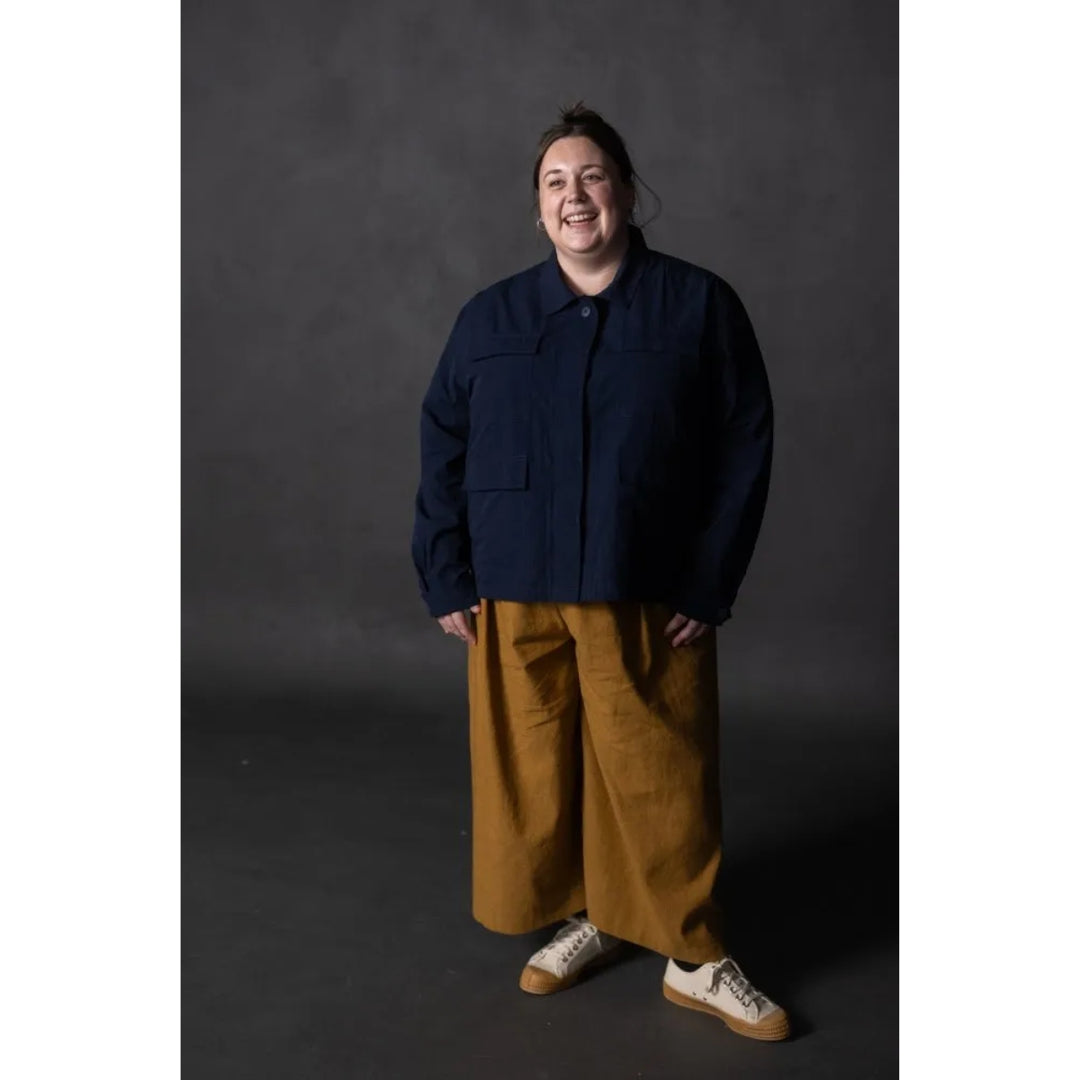 A person standing against a dark gray studio background, wearing a dark navy cropped jacket, wide-leg mustard yellow pants, and white sneakers with tan soles.