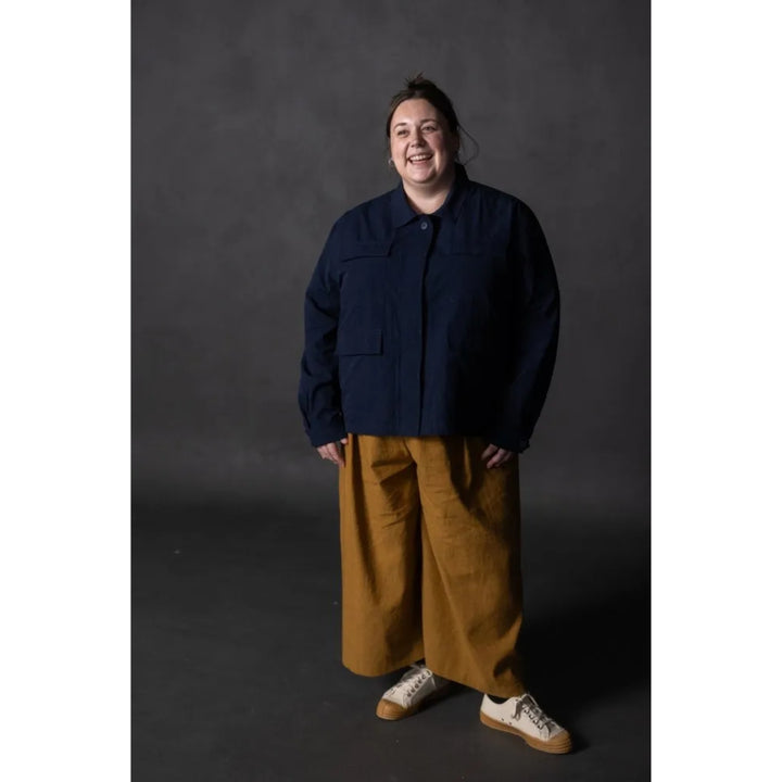 A person standing against a dark gray studio background, wearing a dark navy cropped jacket, wide-leg mustard yellow pants, and white sneakers with tan soles.