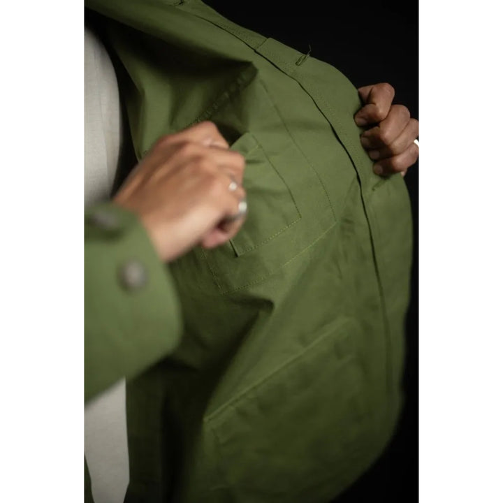 Close-up of a person wearing an olive green jacket, holding the lapels open to show the interior lining and pocket details against a dark background.