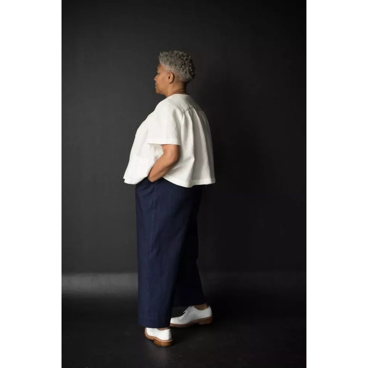 A person standing in profile against a dark studio background, wearing a loose white short-sleeve top, dark navy wide-leg pants, and white shoes.