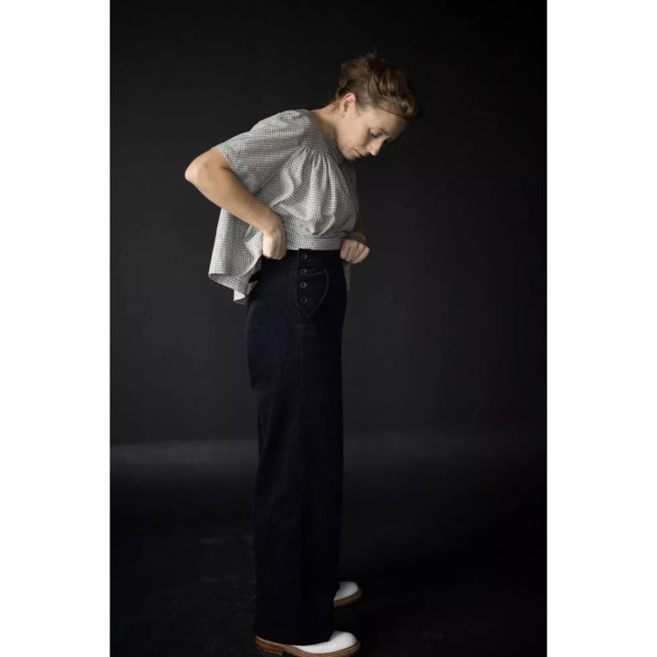 A model wearing a light gray patterned short-sleeve top and dark wide-leg jeans, standing against a dark studio background.