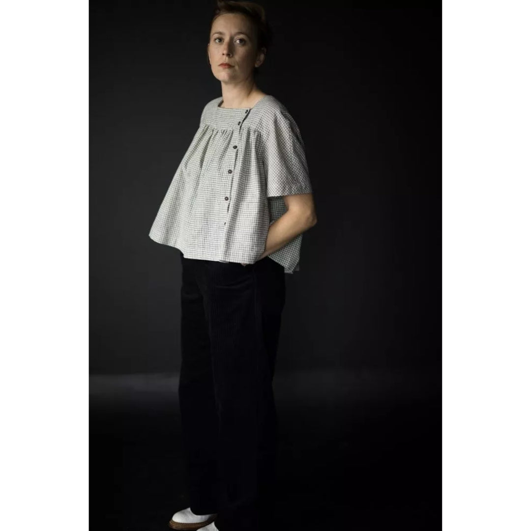 A woman modeling a short-sleeve top with a fine black and white gingham pattern, featuring a gathered neckline and front buttons, paired with black trousers, set against a dark studio background.