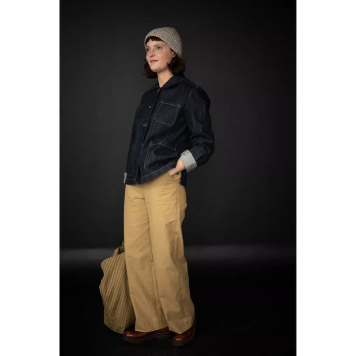 A model wearing a dark indigo denim jacket with rolled-up sleeves, beige wide-leg cargo pants, a gray knit beanie, and brown leather shoes, standing against a dark studio background.