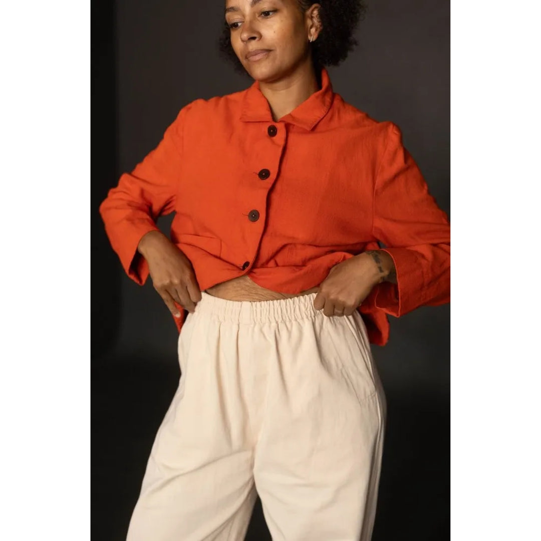 A model wearing a bright orange cropped button-up shirt and cream-colored trousers, standing against a dark studio background.