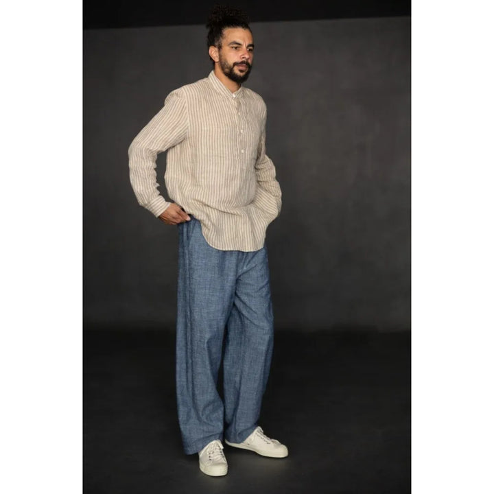 A man modeling a beige and white vertically striped long-sleeve button-up shirt with wide-leg blue jeans and white sneakers, standing against a dark gray studio background.