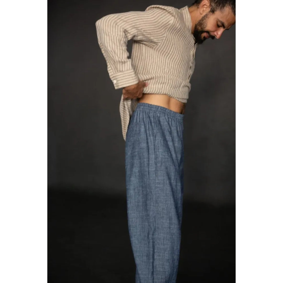 A man wearing a beige and white vertically striped cropped long-sleeve shirt and blue relaxed-fit pants, shown from the waist up against a dark studio background.