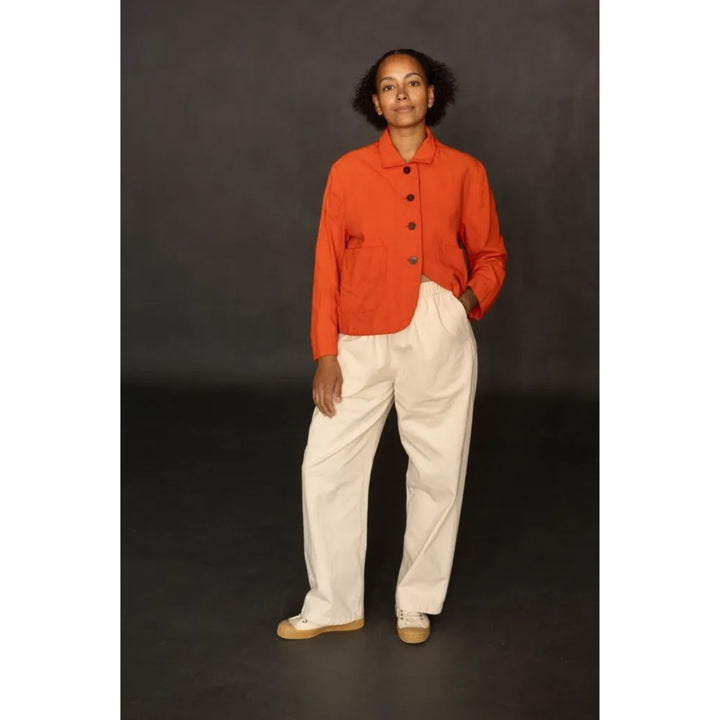 A model stands against a dark gray studio background wearing a bright orange cropped button-up shirt and off-white wide-leg trousers.