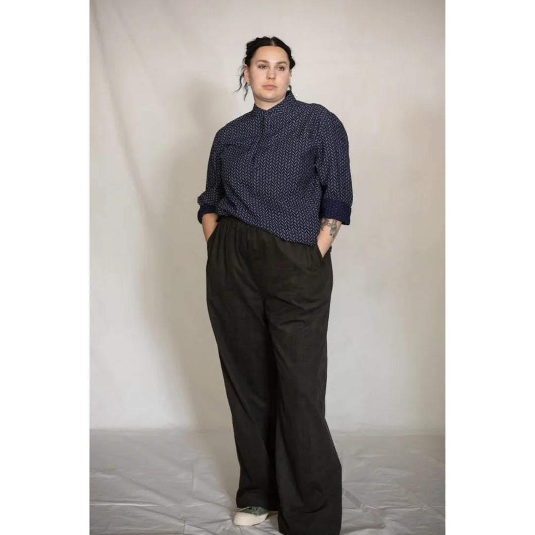 A model stands against a light beige studio backdrop wearing a dark navy blue high-neck button-up shirt with a small white geometric pattern, paired with loose black wide-leg trousers and white sneakers.