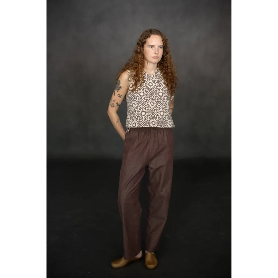 A model with long curly brown hair stands against a dark gray studio backdrop, wearing a sleeveless cream top with a brown geometric pattern and loose brown pants.
