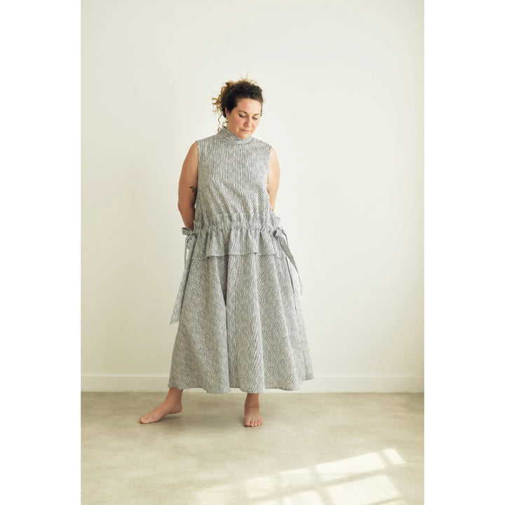 A woman wearing a sleeveless, high-neck, tiered midi dress with a gray and white textured pattern, standing barefoot in a minimalist room with a cream wall and concrete floor.