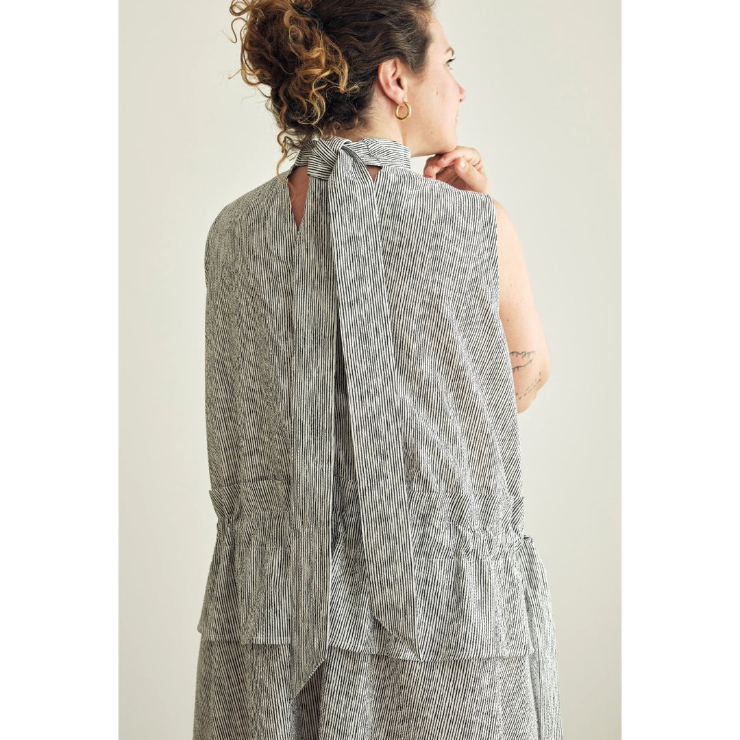 A woman shown from the back wearing a sleeveless gray and white vertically striped top with a tie-neck detail and layered ruffle hem, against a plain light beige background.