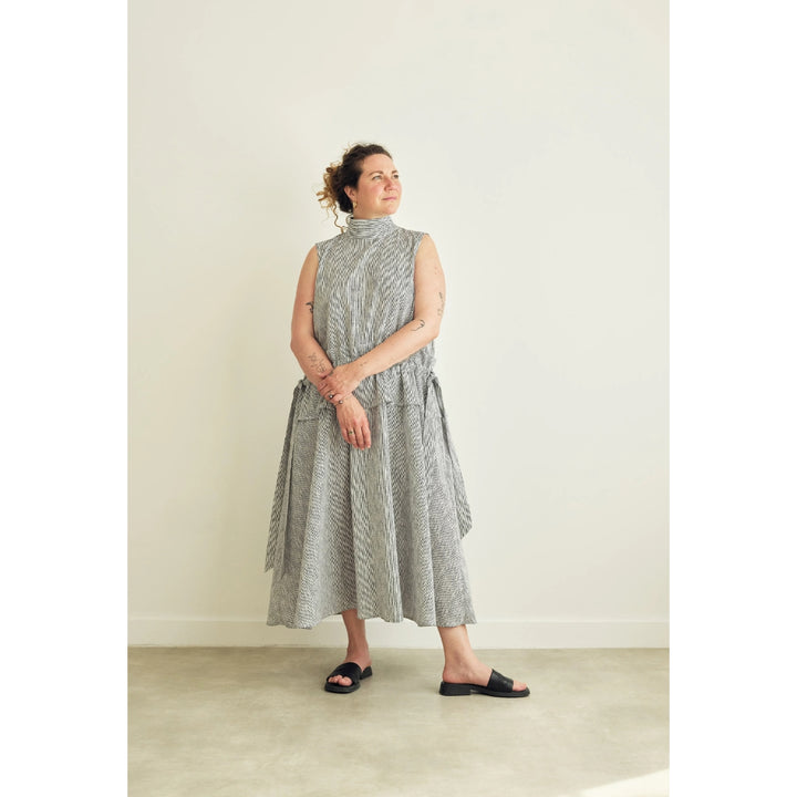 A woman wearing a sleeveless, high-neck midi dress with a textured gray and white pattern, standing in a minimalist room with a light beige wall and concrete floor.