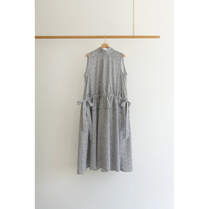 A sleeveless gray textured dress with a high mock neck, gathered waist, and side tie details, hanging on a wooden hanger from a horizontal wooden rod against a plain white wall.