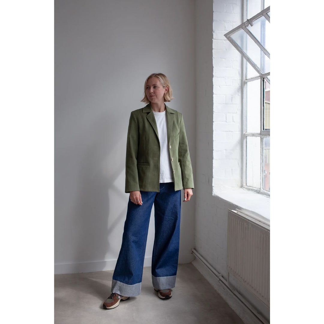 Person wearing a green jacket and blue jeans standing in a room with a large window.