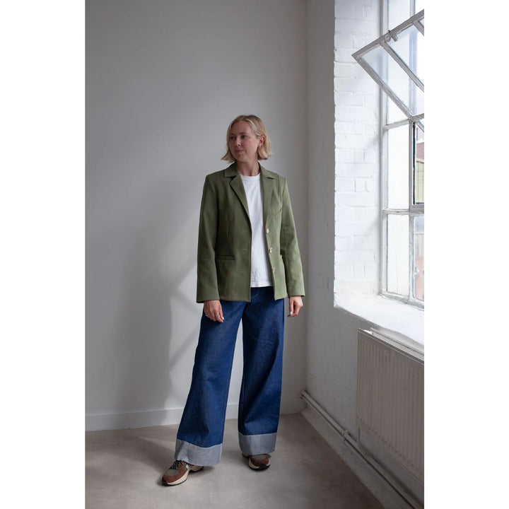 Person wearing a green jacket and blue jeans standing in a room with a large window.