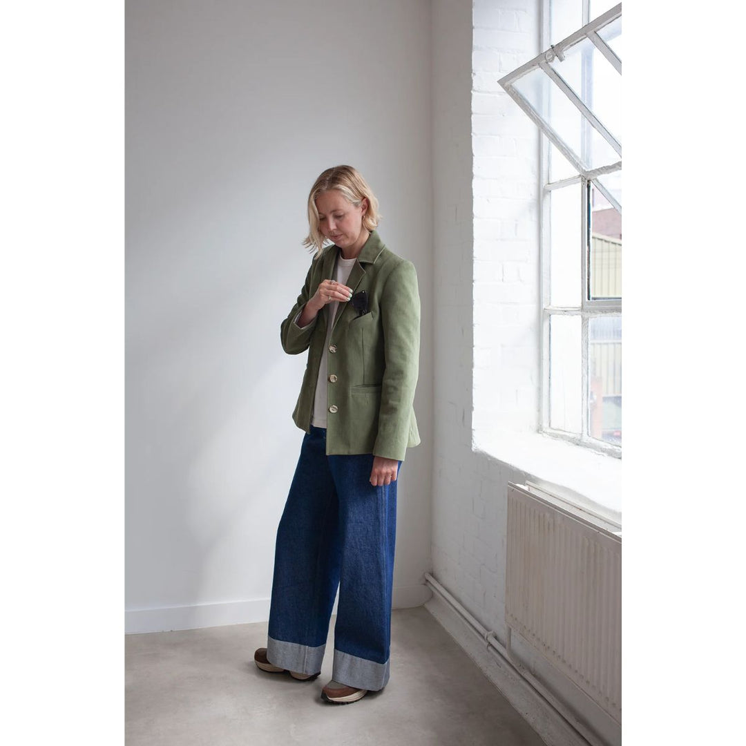 Woman in a green jacket and blue pants standing in a room with large windows.