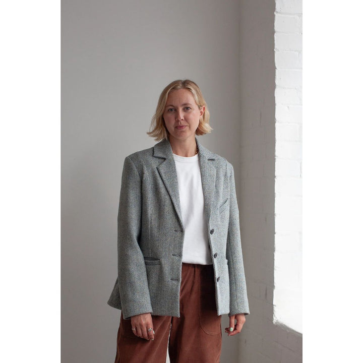 Woman wearing a gray blazer over a white shirt and brown pants against a plain wall.
