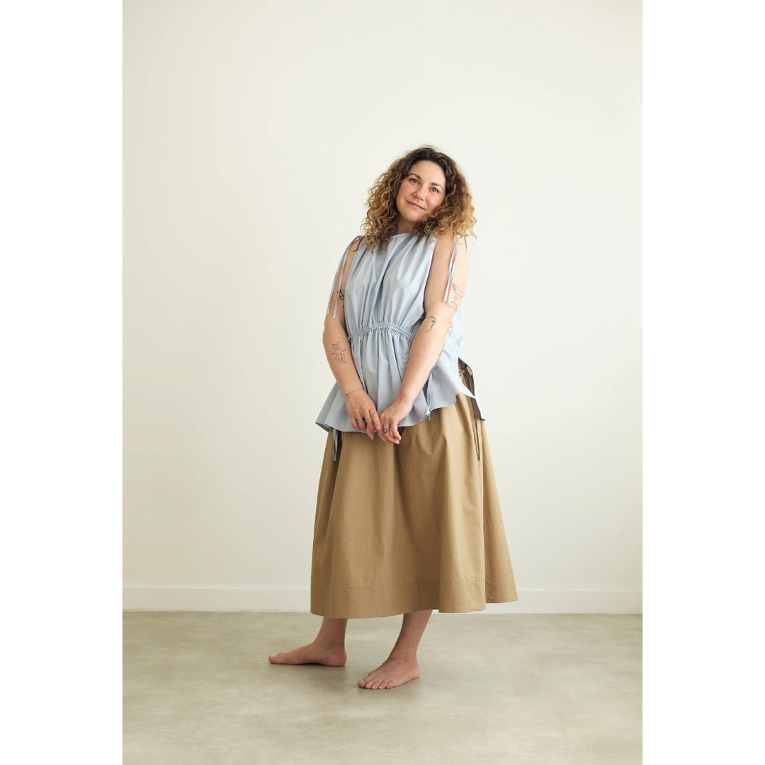 A woman stands barefoot in a minimalist studio setting, wearing a light blue sleeveless top with a gathered waist and a long tan skirt.