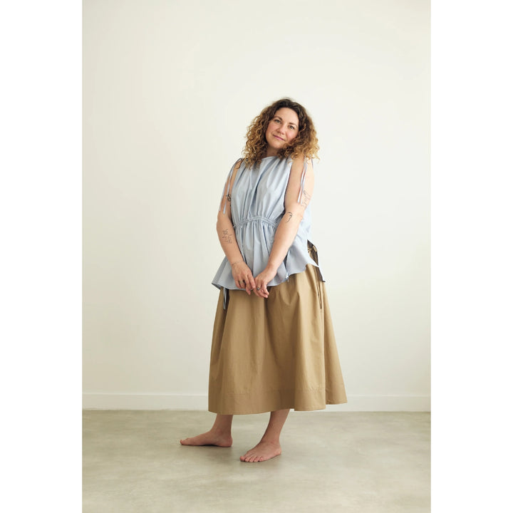 A woman stands barefoot in a minimalist studio setting, wearing a light blue sleeveless top with a gathered waist and a long tan skirt.