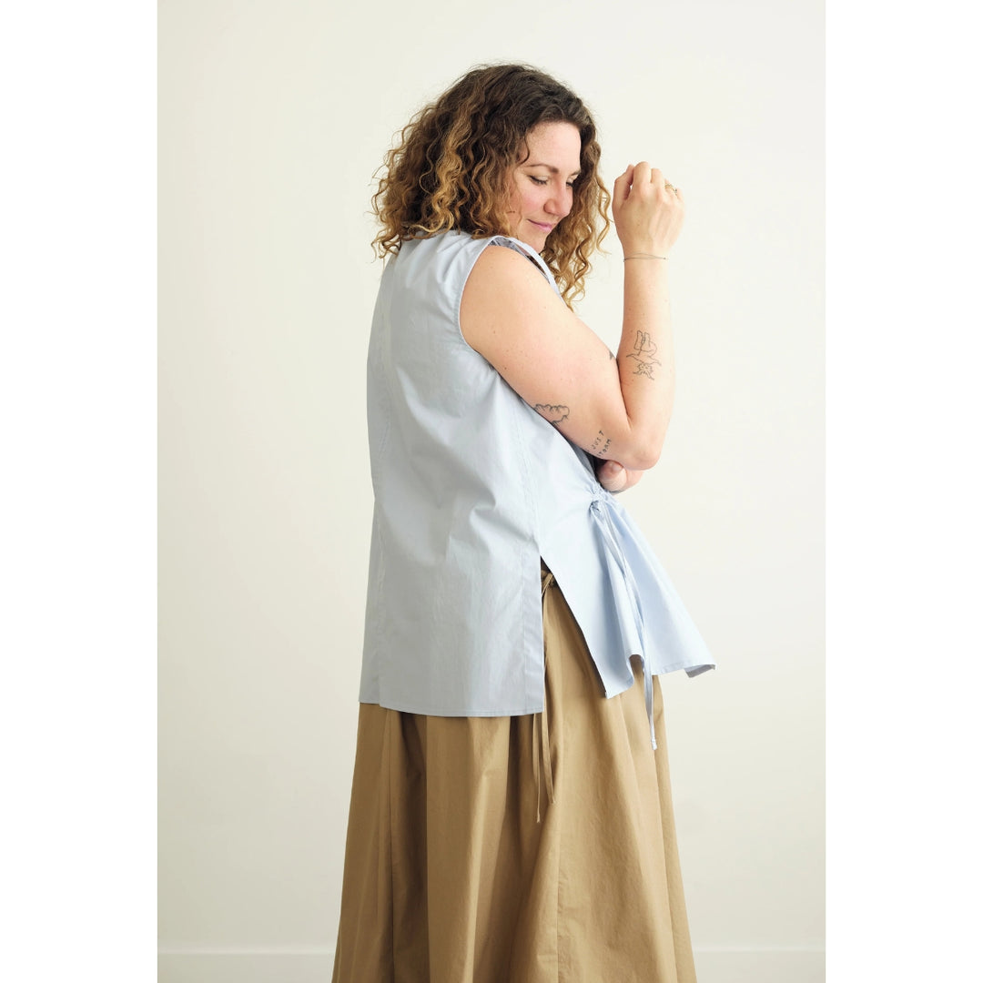 A woman with curly brown hair is shown from the side, wearing a light blue sleeveless wrap-style top and a tan maxi skirt against a plain off-white background.