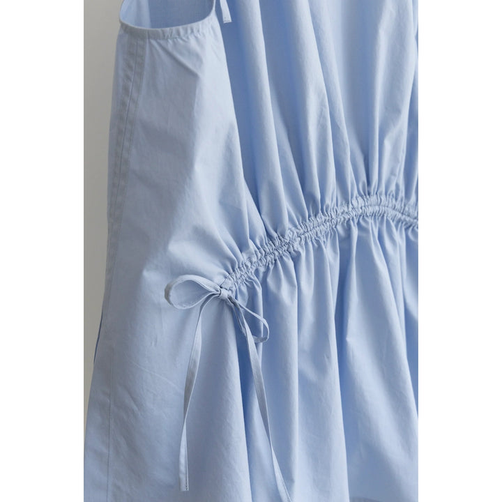 Close-up of a light blue sleeveless top or dress with a gathered elastic waist and a side tie detail, shown against a plain light gray background.