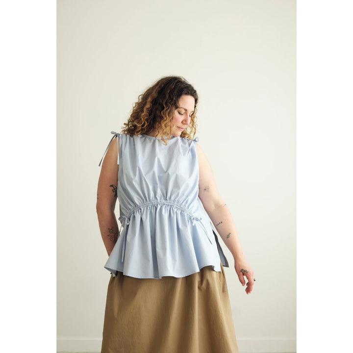 A woman wearing a light blue sleeveless peplum top with tie details at the shoulders and a gathered waist, paired with a brown skirt, standing against a plain off-white wall.