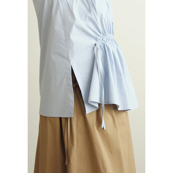 A close-up of a light blue wrap-style blouse with a gathered side detail and drawstring, worn over a tan skirt or pants against a plain light beige background.